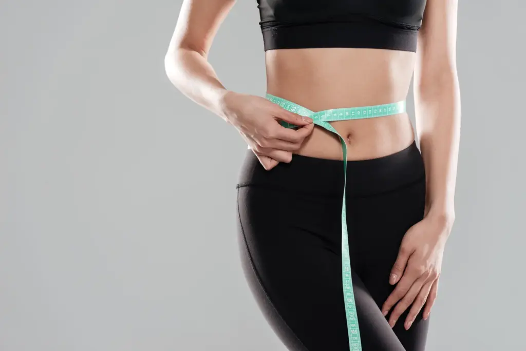 A woman in activewear wraps a tape measure around her waist to track body contouring progress over time.