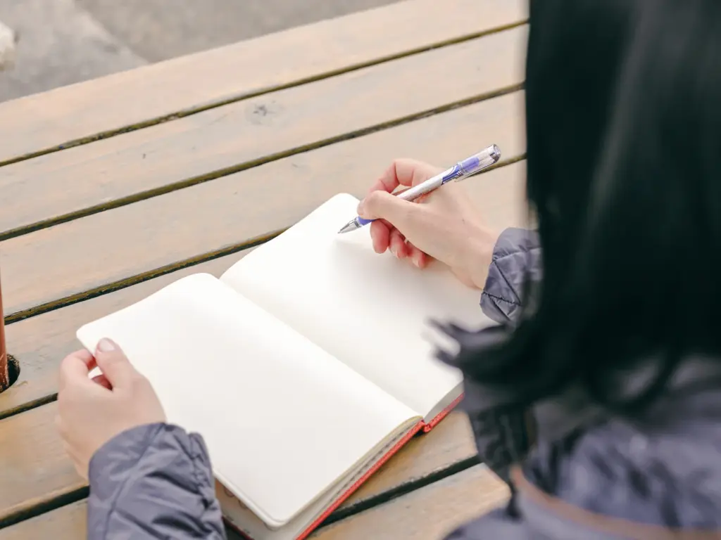 Woman writing in a notebook at an outdoor table, keeping a simple journal of sleep, mood, and cycle changes in her early forties.