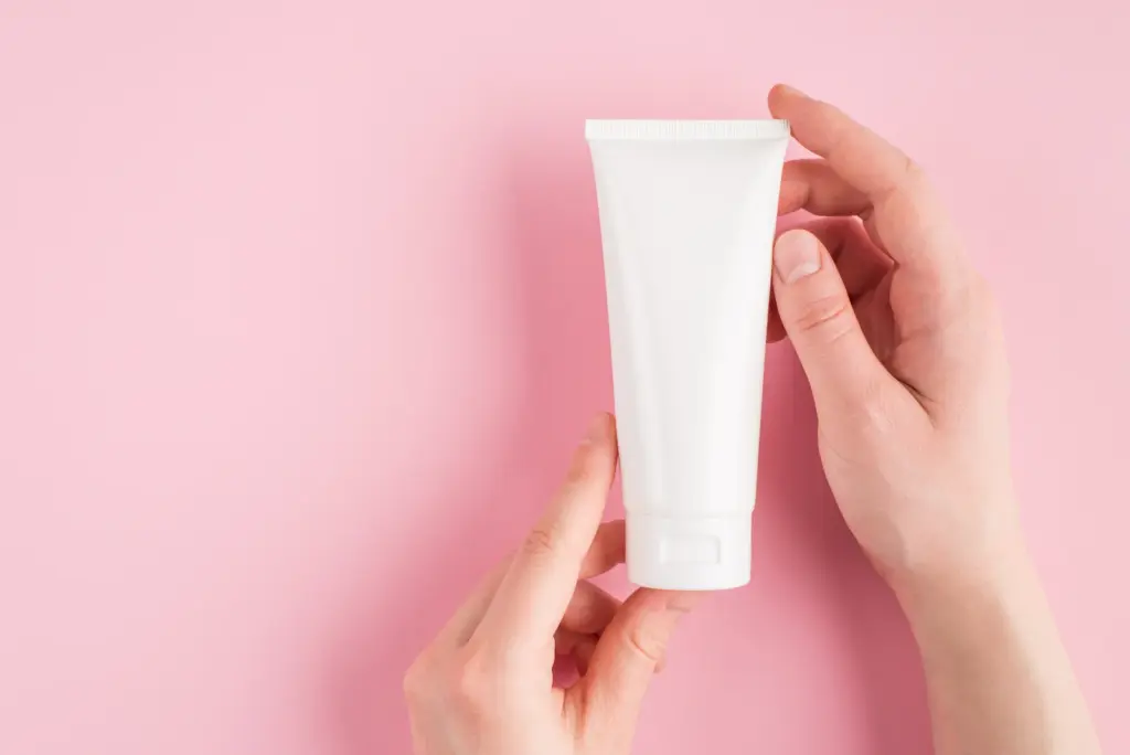 Hands holding a plain white tube of moisturizer against a soft pink background, part of a simple home routine for dry skin in Tempe.