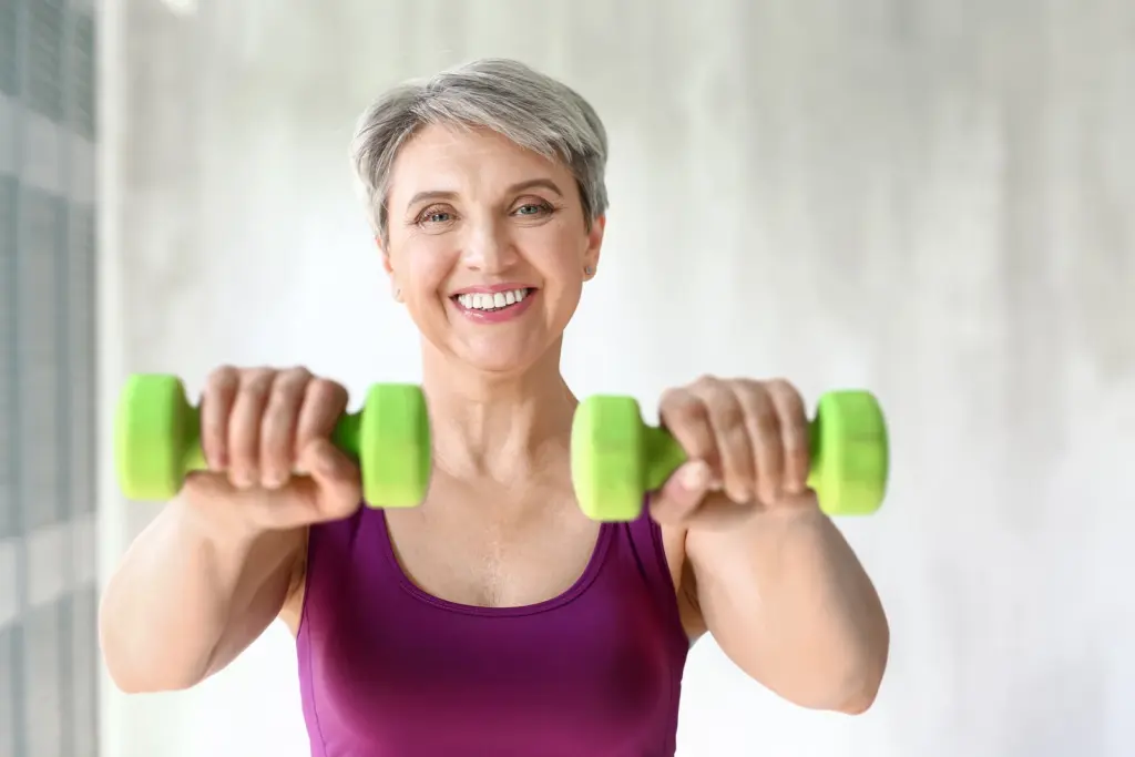 Smiling woman with gray hair lifting light dumbbells at home, strength training that helps protect muscle during menopause in Tempe.