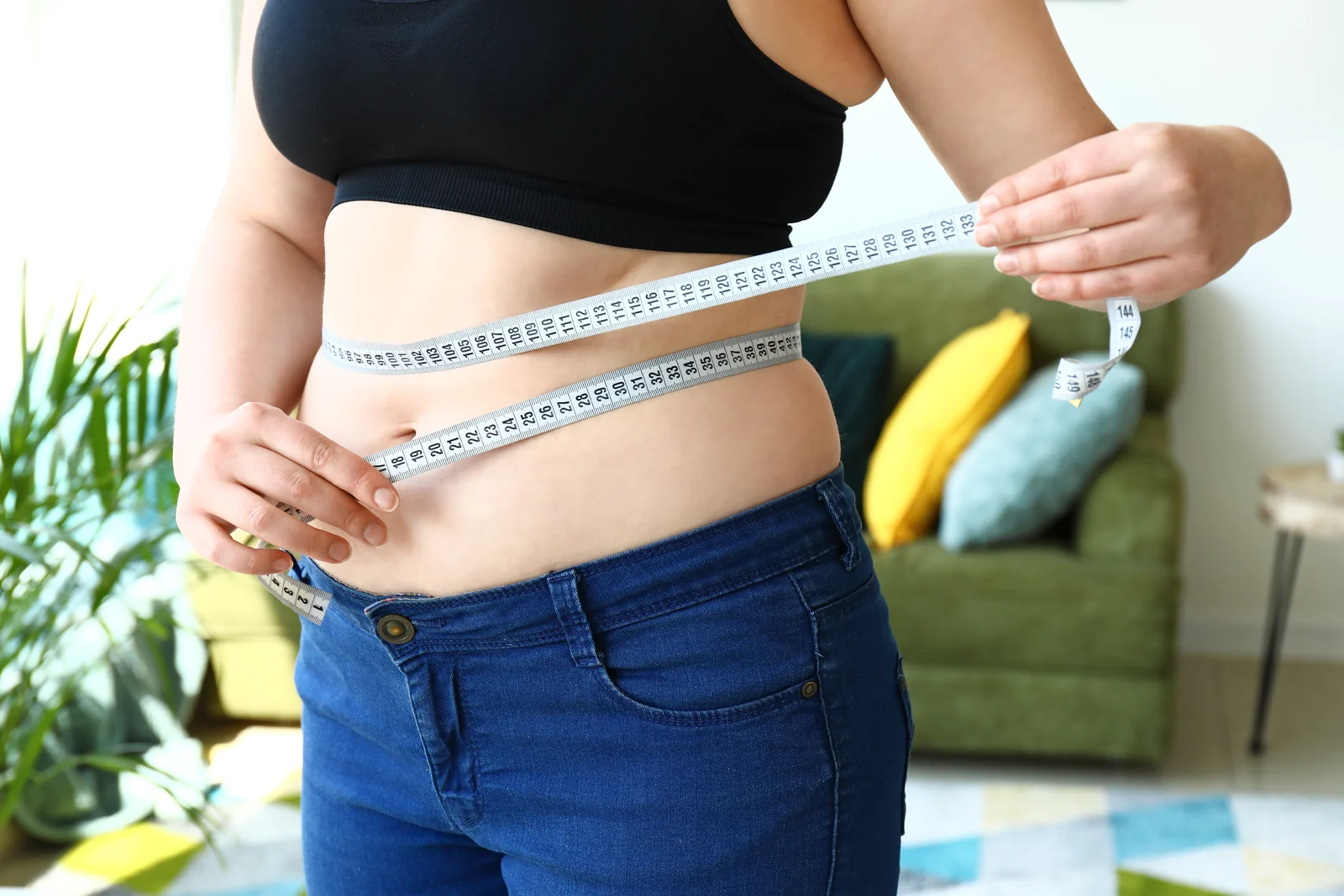 Woman wrapping a measuring tape around her waist at home, tracking the belly weight many women notice during menopause in South Tempe, AZ.