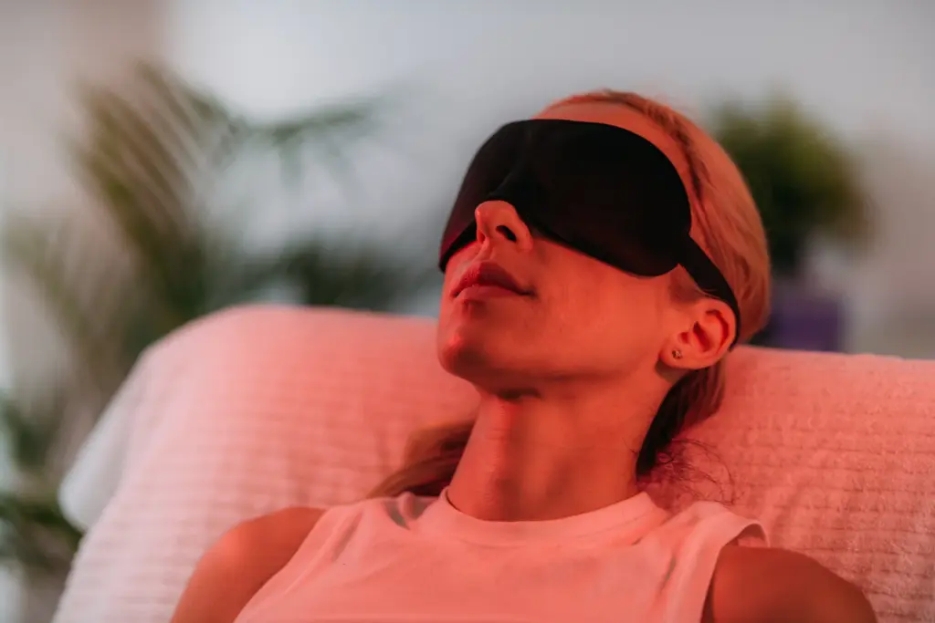 Woman resting with an eye mask under a warm red light therapy lamp, the low-level light approach used in some hair loss plans.
