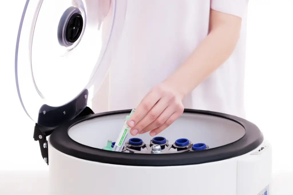 Gloved hand placing a blood sample tube into a centrifuge, the step that separates platelet-rich plasma for PRP treatments.