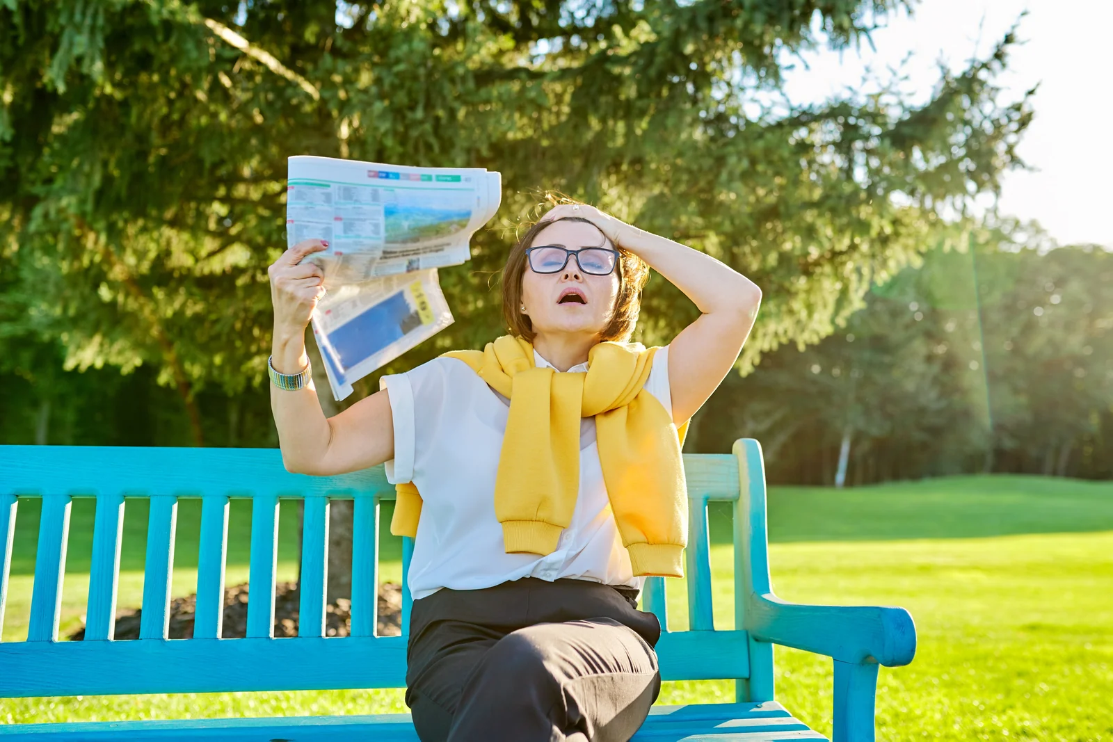 Woman fanning herself with a newspaper on a park bench during a hot flash, a common perimenopause symptom for women in South Tempe, AZ.